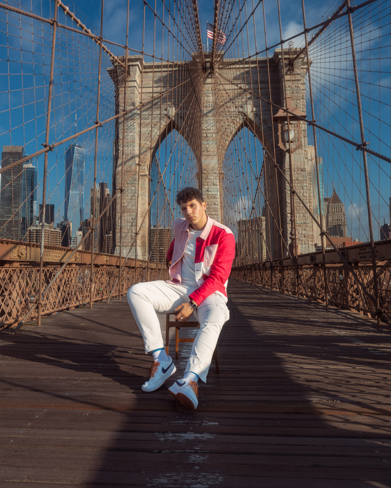 Wide portrait of ILIJA seated on the Brooklyn Bridge walkway in a red and pink jacket