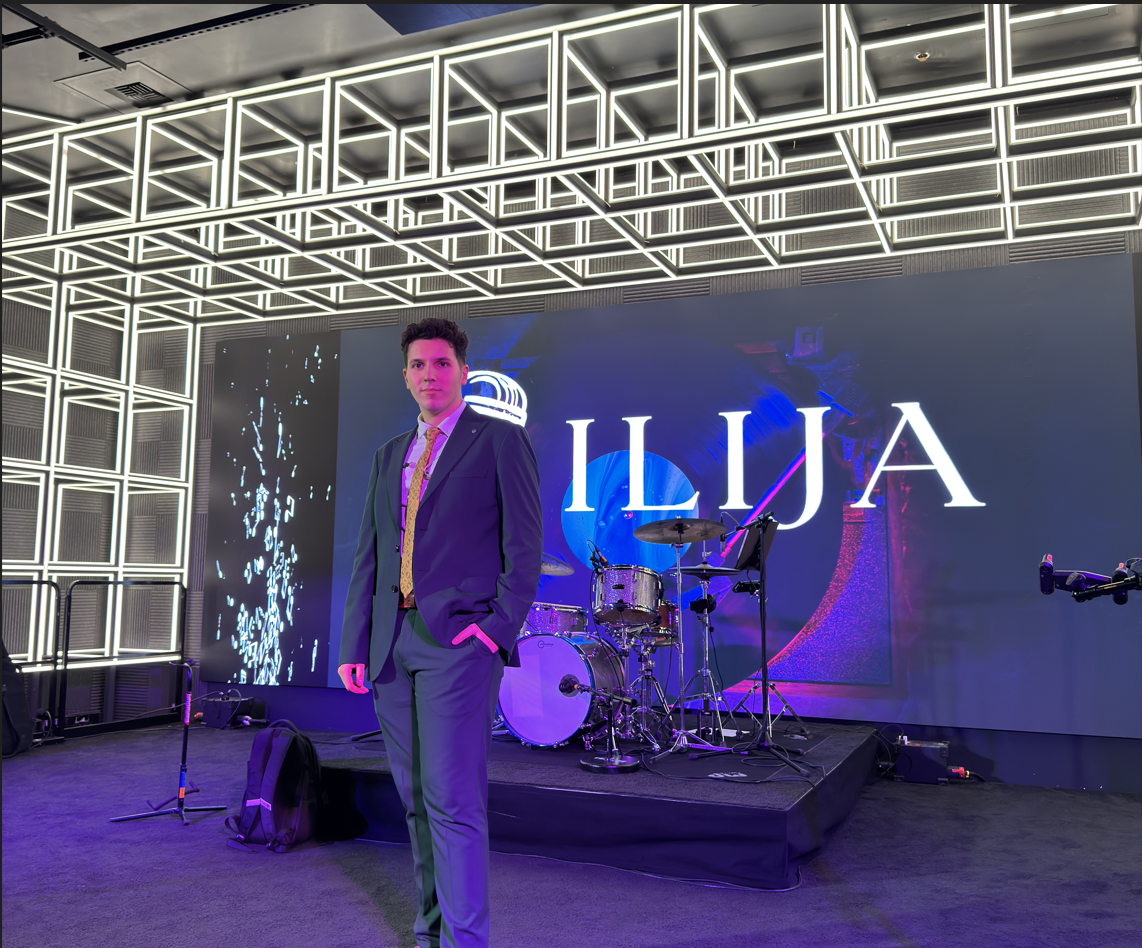 ILIJA on stage in a suit and gold tie in front of a large neon ILIJA sign and drum kit