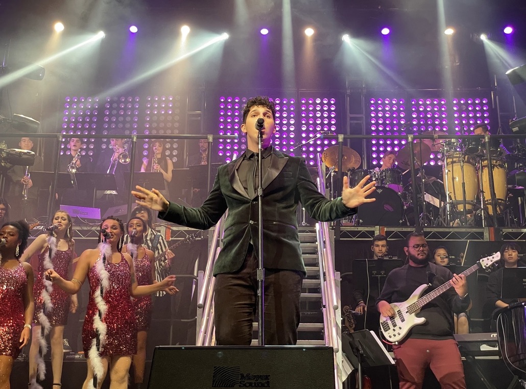 ILIJA singing lead with a full big band and backing vocalists under purple stage lights