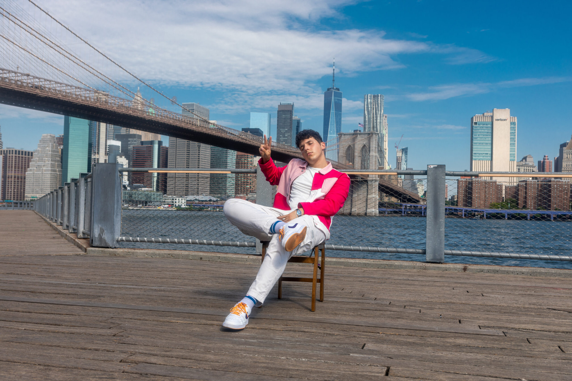 ILIJA seated on a chair on the Dumbo waterfront with the Brooklyn Bridge and Manhattan skyline behind him