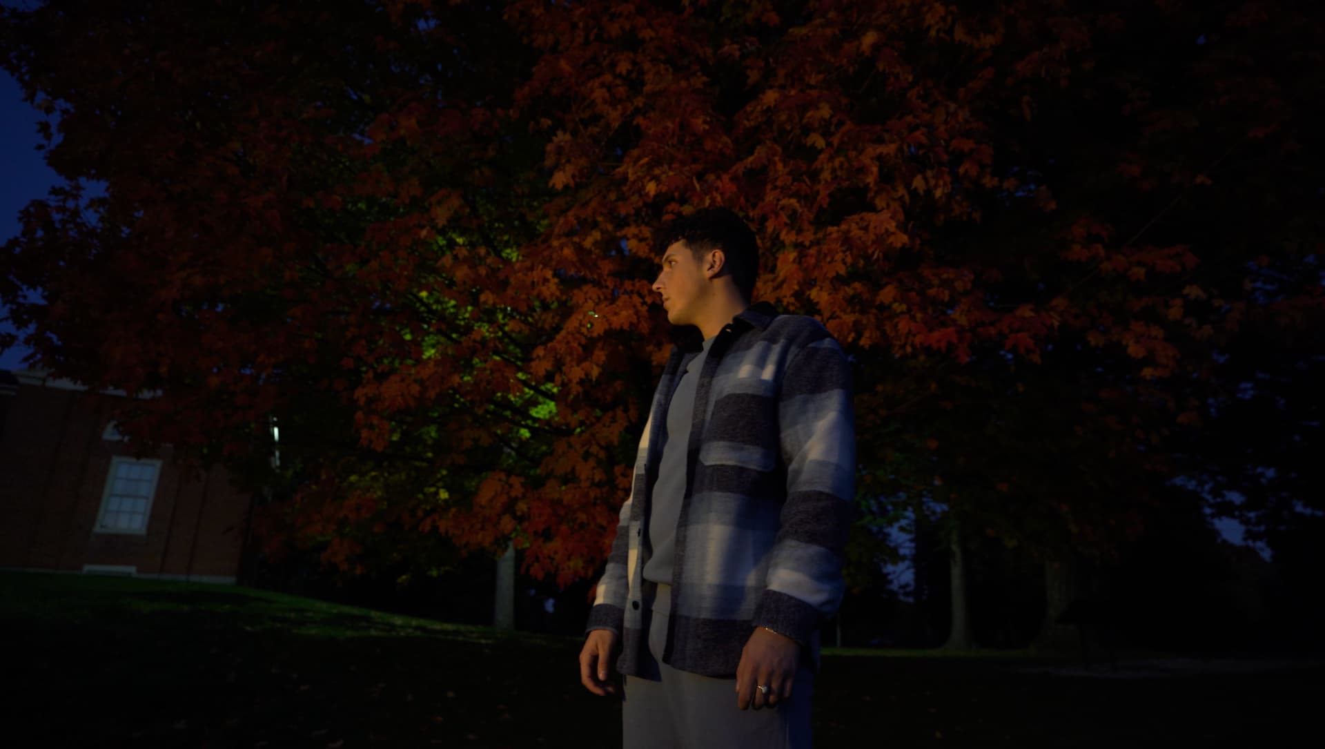 ILIJA outdoors at twilight in front of a tree with vibrant red and orange autumn leaves, wearing a blue and white plaid jacket