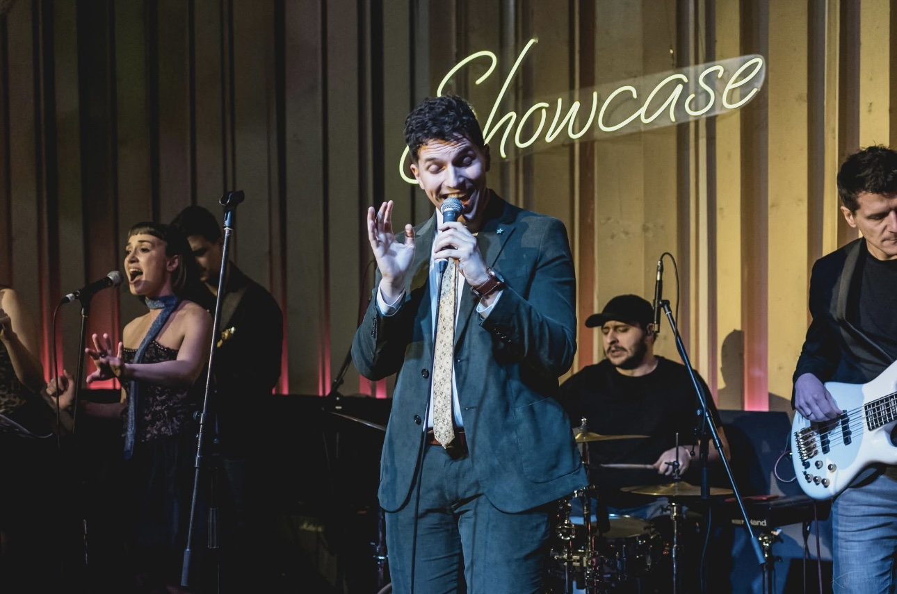 ILIJA singing into a handheld microphone with backing vocalists at the Showcase performance