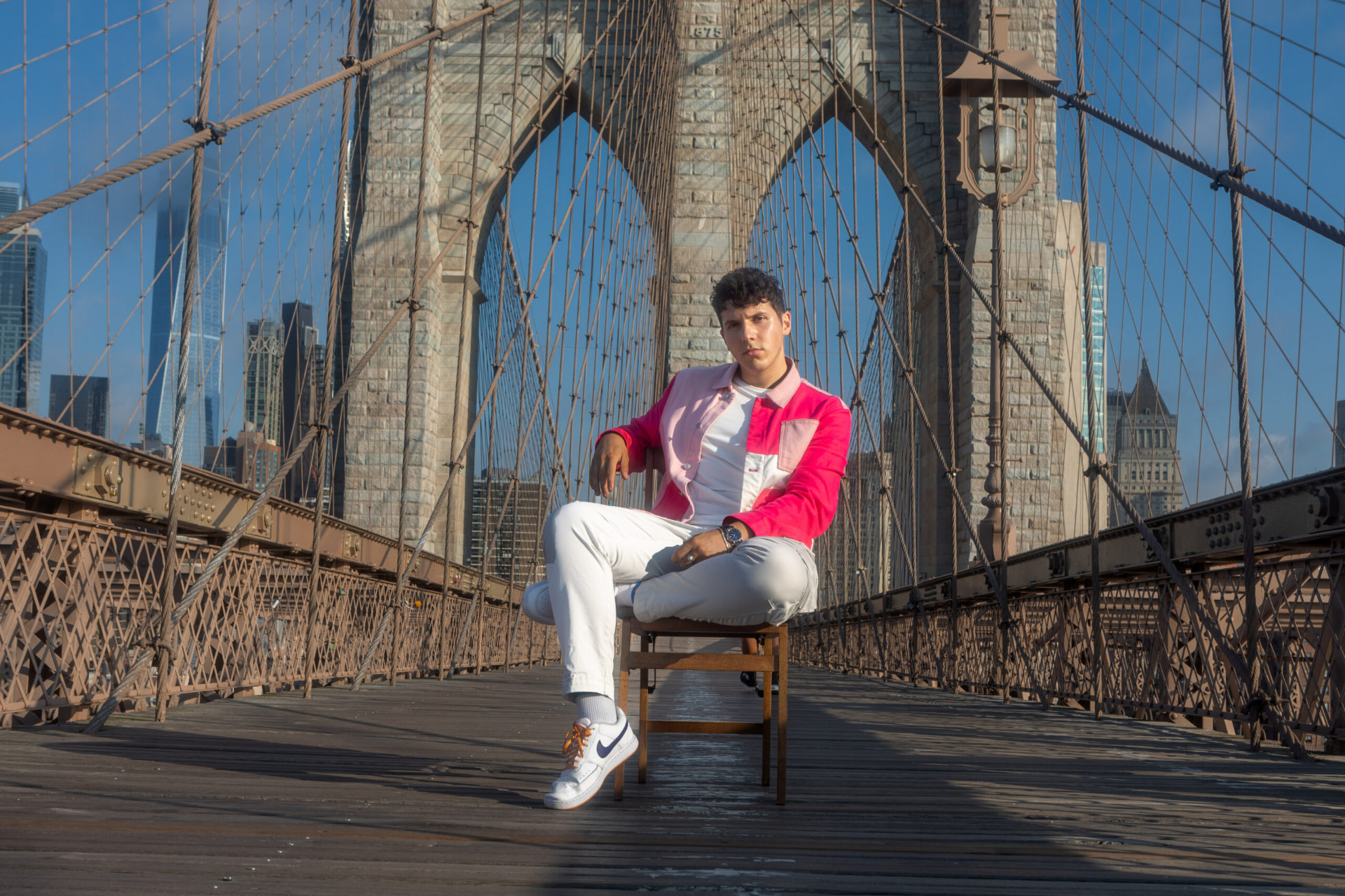 ILIJA looking into the camera while seated on a wooden chair on the Brooklyn Bridge