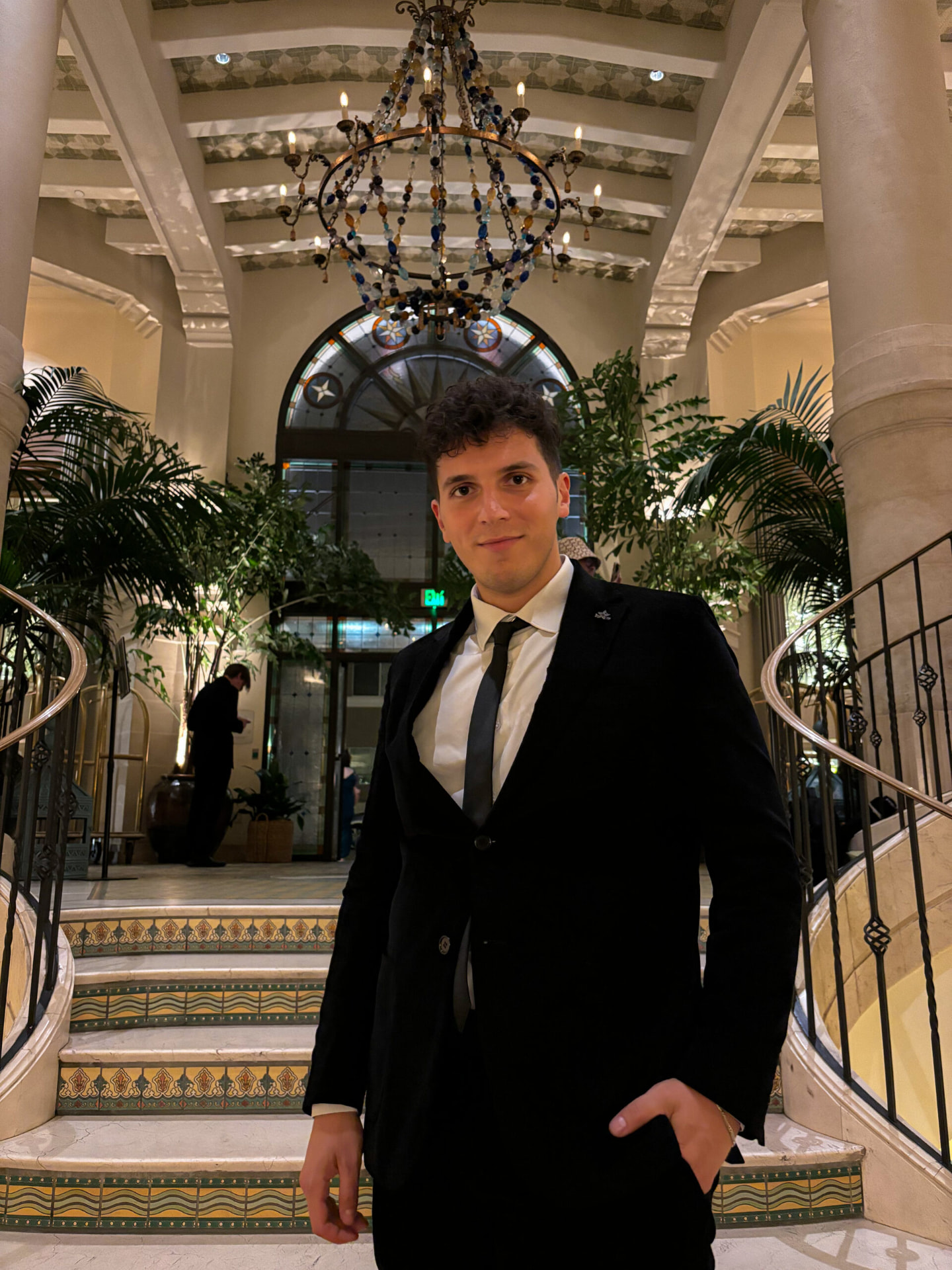 ILIJA in a black suit and tie on a grand hotel staircase beneath a chandelier