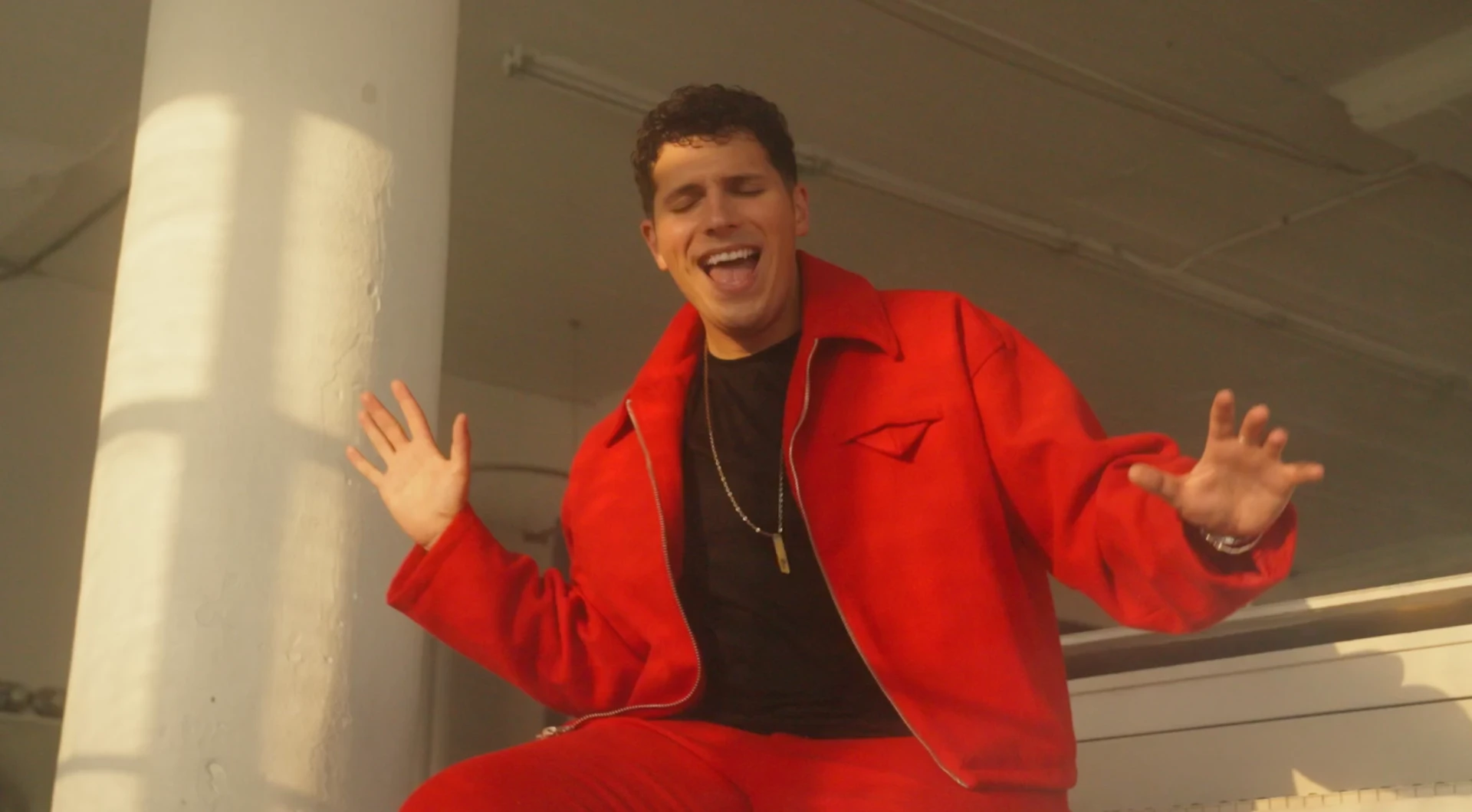ILIJA in a bright red jacket and trousers singing on top of a white piano