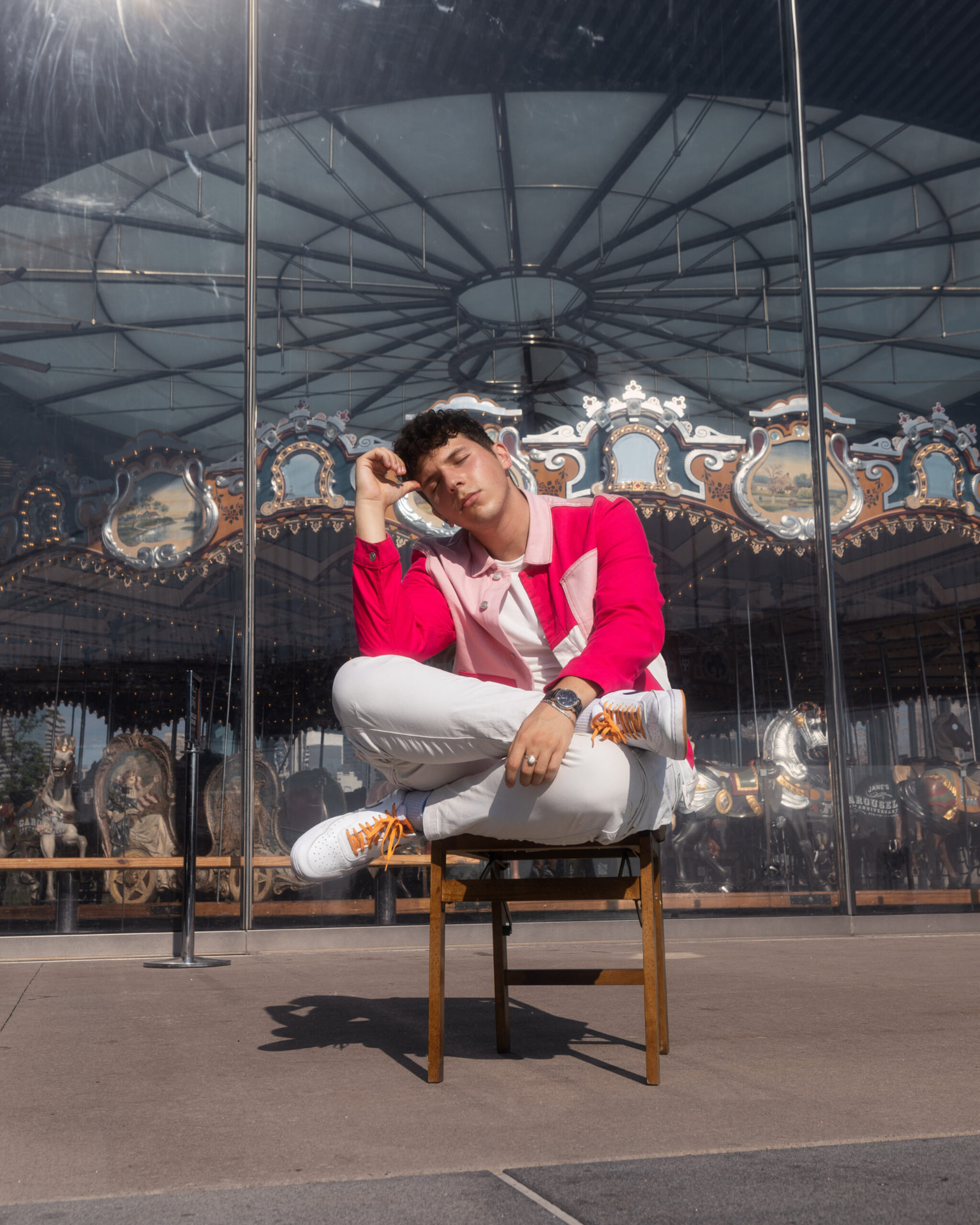 ILIJA sitting cross-legged on a chair in front of Jane's Carousel in Brooklyn