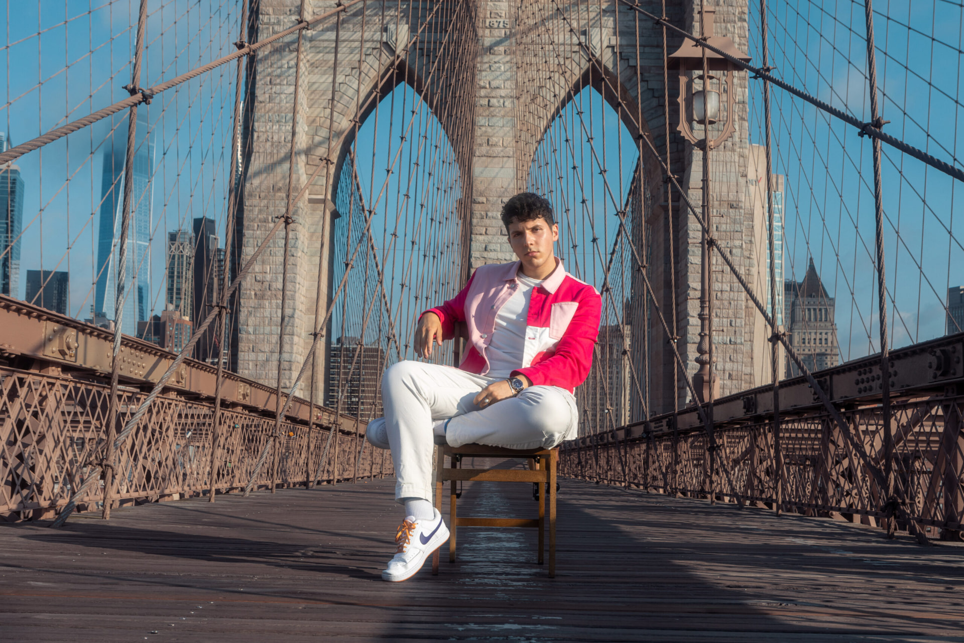 ILIJA seated with legs crossed on the Brooklyn Bridge, downtown Manhattan behind him