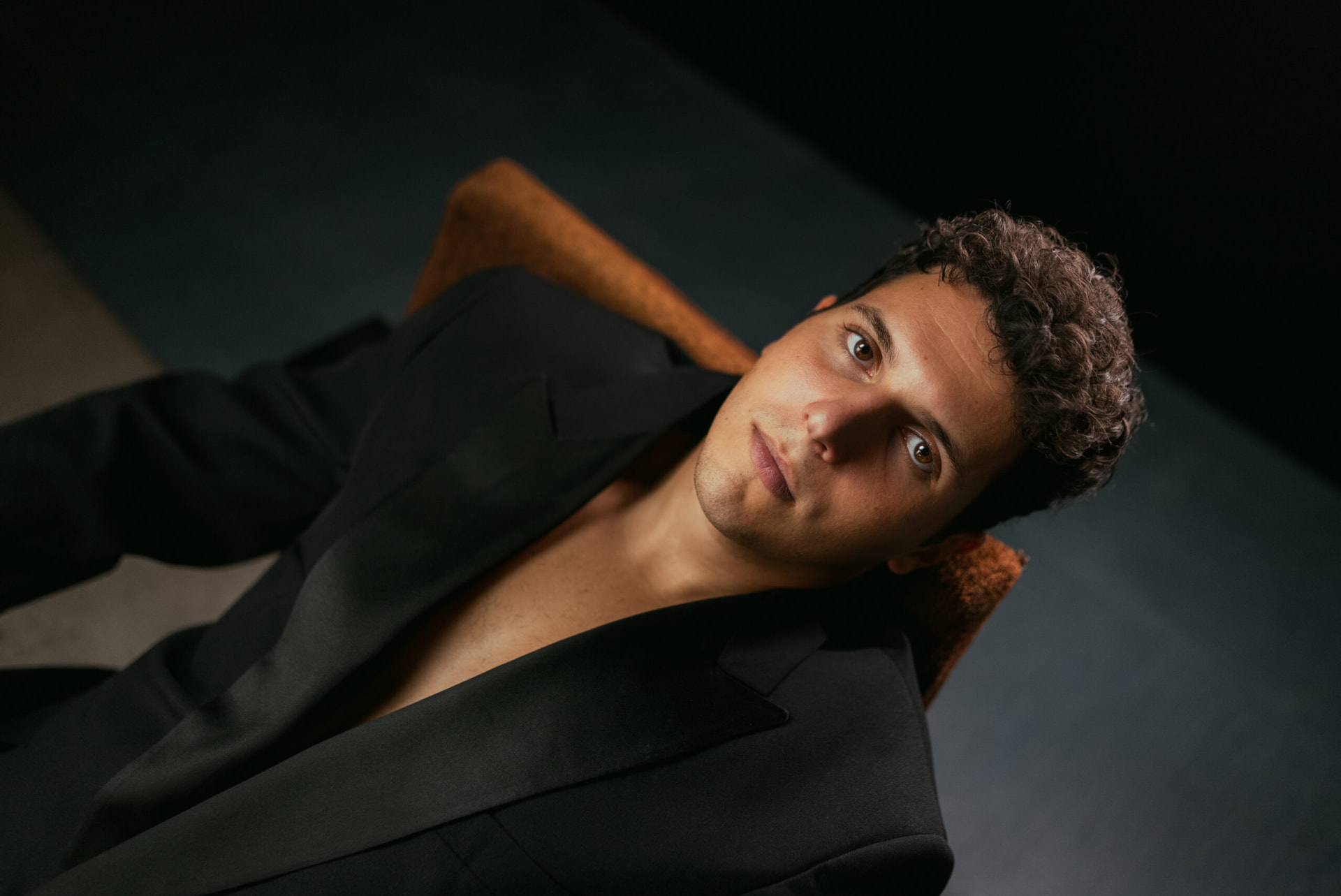 Moody studio portrait of ILIJA reclining in a black suit against a dark backdrop